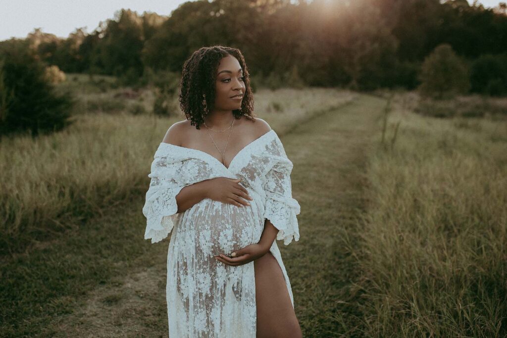 Maternity Photo Session in Fayetteville NC by Victoria Vasilyeva Photography. This photo shows a pregnant mom posing among high grass. She is wearing a white lace dress, and the sun is shining through the leaves, creating a beautiful halo effect around her. The photo is a timeless reminder of this special time in her life.