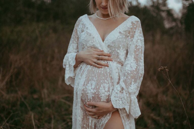The photo of tiny beautiful pregnant belly. Mom-to-be is holding it and looking down. She wears lace white maxi dress. The portrait session was done by Victoria Vasilyeva Photography.