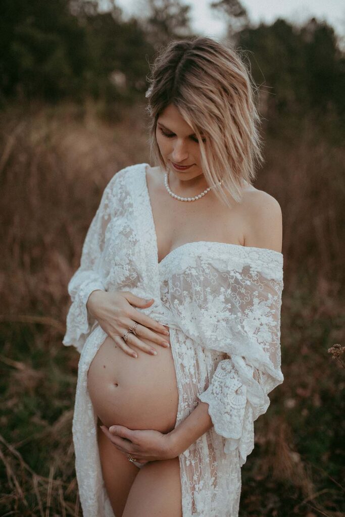 Gorgeous mom-to-be in maxi white dress among high grass. She holds her belly and looks down at it. The maternty portrait was taken by Victoria Vasilyeva Photography near baby shower venues Greensboro NC.