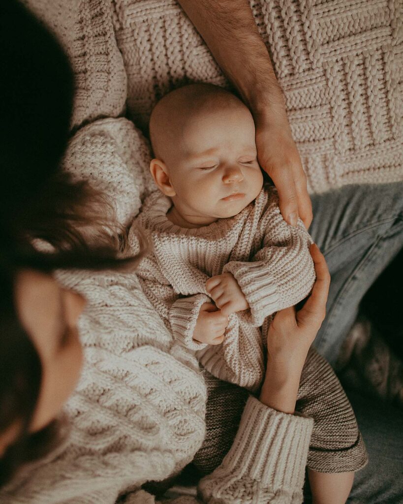 Cute baby girl in tan knitted swetaer is peacfully sleeping under the Christmas tree in her mom's lap. It was bought at one of the Christmas tree farms near Raleigh NC. Victoria Vasilyeva Photography - Raleigh Family Photographer.