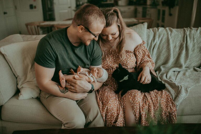 A playful black cat, adorned with an elegant bow, adds a touch of whimsy to the scene, captivating the attention of the newborn baby girl. Captured by Clayton Newborn Photographer - Victoria Vasilyeva Photography.