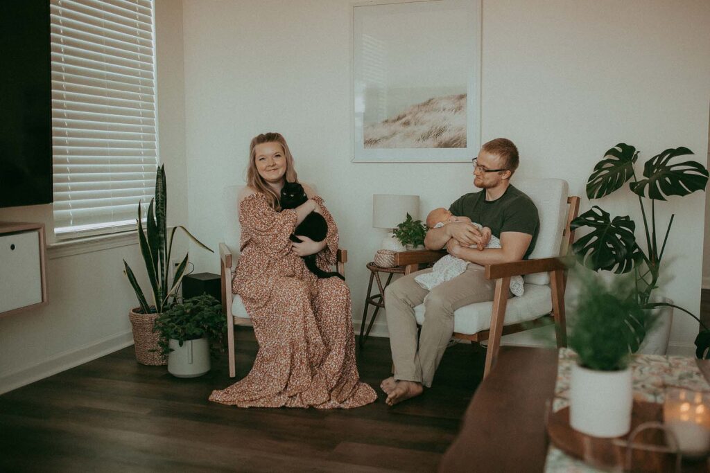 A loving family portrait, captured amidst the vibrant greenery of their home, showcases the unbreakable bond between a mother, father, newborn daughter, and their feline companion. Captured by Clayton Newborn Photographer - Victoria Vasilyeva Photography.