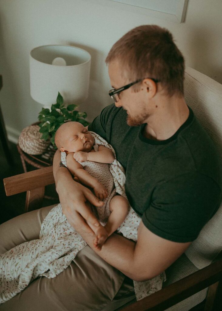 A father protectively embraces his newborn daughter, his green t-shirt and tan pants blending seamlessly with the earthy tones of their home, creating a sense of comfort and security. Captured by Clayton Newborn Photographer - Victoria Vasilyeva Photography.