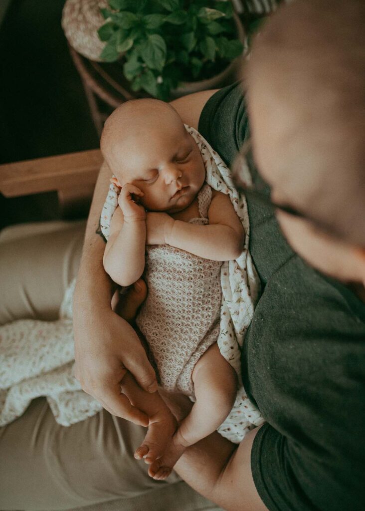 A father's heart overflows with love as he gazes adoringly at his newborn daughter, his green t-shirt and tan pants complementing the serene atmosphere. Captured by Clayton Newborn Photographer - Victoria Vasilyeva Photography.
