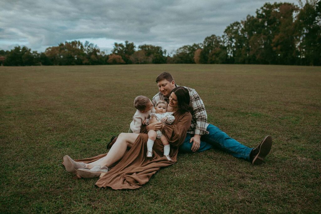 Family of four is sitting on a field. Dad hugs mom and mom holds her daughter. They look happy and enjoy each other. The photo was taken at Lake Benson Park by Victoria Vasilyeva Photographer.