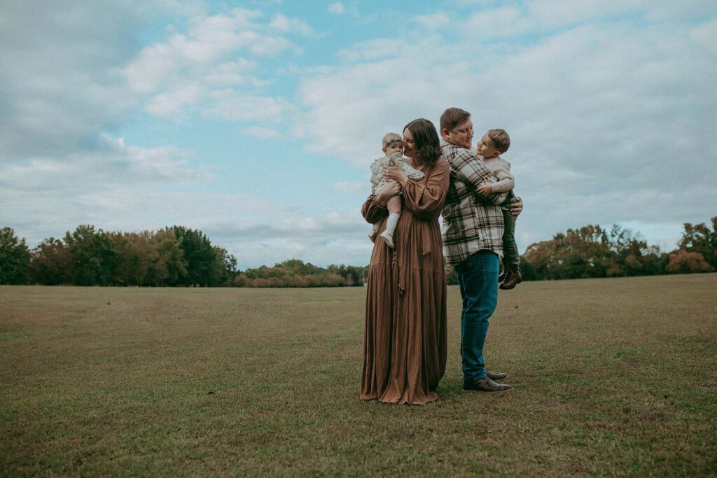 Family of four is standing on a field. Dad holds his son and mom holds her daughter. They look happy and enjoy each other. The photo was taken at Lake Benson Park by Victoria Vasilyeva Photographer.