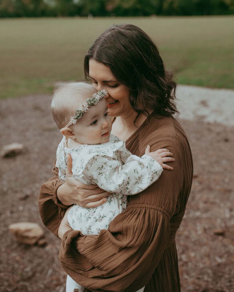 Mom in boho brown dress is holding her baby girl and looking down at her. Baby girl is smiling and looking at her right. She wear beautiful white romper and flower halo. The photo was taken near Clayton daycare by Victoria Vasilyeva Photographer.