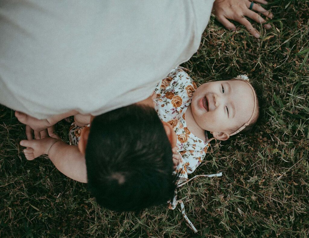 Cute and adorable baby girl in floral romper lays on a grass and laughs. Her dad tickles her belly. Photo session took place near daycare in Chapel Hill with family photographer Victoria Vasilyeva Photography.