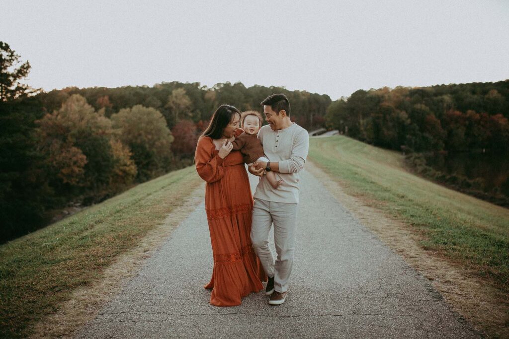 Mom, Dad and 10 months old baby girl walk along the road and smile to each other. Mom wears boho orange dress and has long black hair. Dad wears grey and white clothes. Baby girl wears knitted brown sweater and white pants. Family photo session by Victoria Vasilyeva Photography.