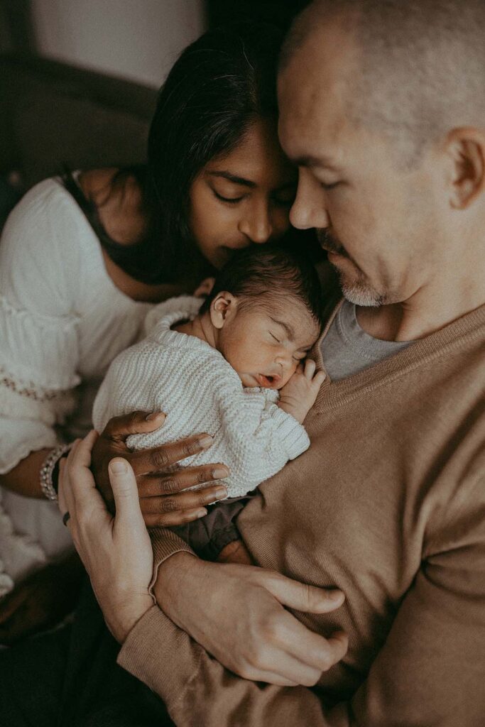 A portrait of mom, dad and newborn baby boy. They are wearing off-white and tan clothes. Baby boy is sleeping on dad's chest. Mom is kissing baby's head natural baby doulas victoria vasilyeva photographer - greensboro newborn photographer.