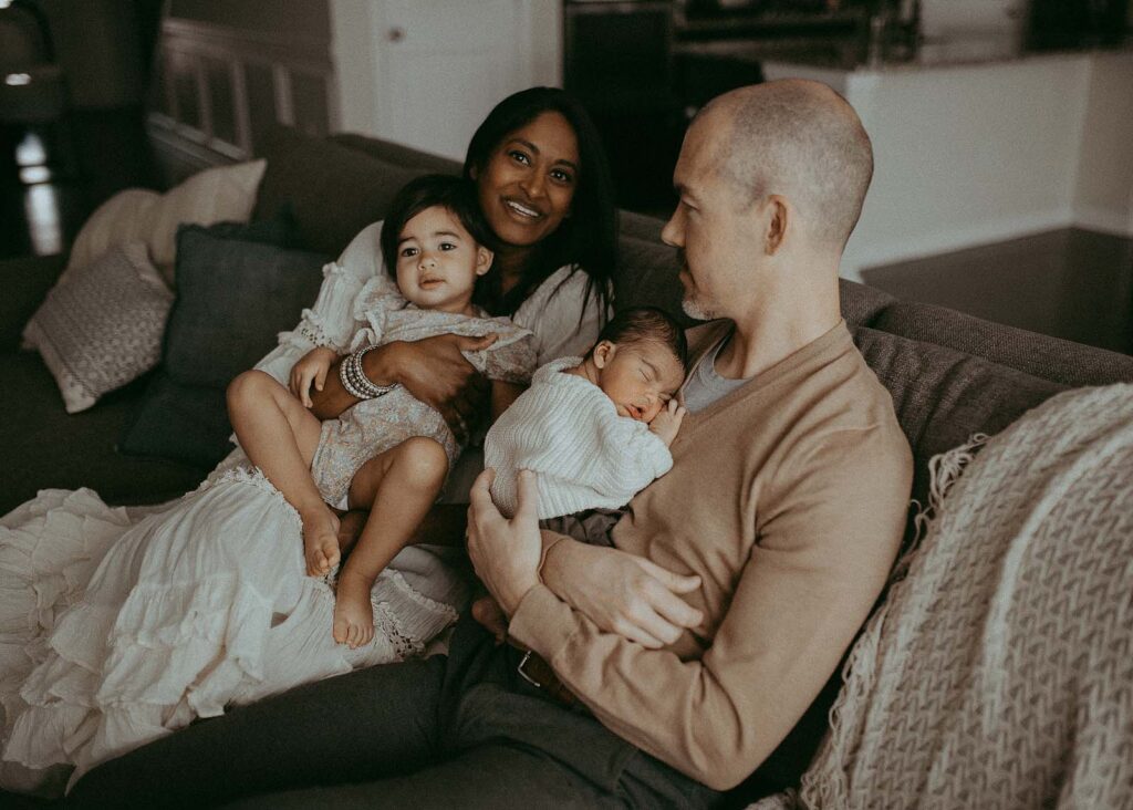 Family of four is wearing boho white and tan clothes is sitting on a sofa in a living room and posing to Greensboro newborn photographer - Victoria Vasilyeva Photographer natural baby doulas.