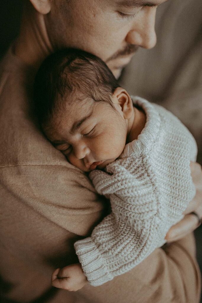 Newborn baby boy with chubby cheeks in cozy knitted sweater from Old Navy is sleeping on dad's shoulder natural baby doulas victoria vasilyeva photographer