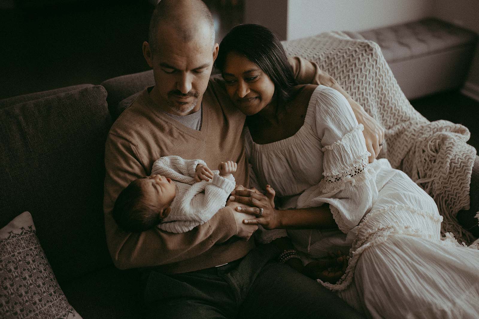 Cozy and beautiful portrait of family of three. Dad is holding a newborn baby and looking at him with love. Mom is sitting newt to him and looking at baby. They are wearing cozy and stylish clothes in earthy tones. The portrait was taken by - victoria vasilyeva photographer - greensboro newborn photographer.