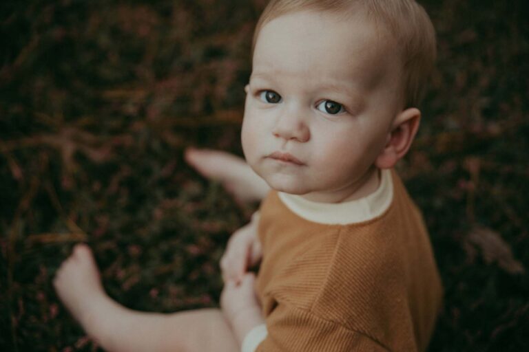 Sweet 10 months old baby boy sits on a grass and looks back. He has brown eyes and short blonde hair. Family photo session by Victoria Vasilyeva Photography took place in Cary, NC near daycare center.