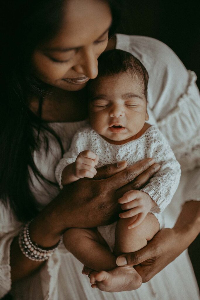 Incredible newborn photoshoot capturing the essence of family love, with a touch of bohemian style. Newborn baby boy is hugged by his mom. They both wear white. Mom smiles and looks at baby. Newborn portrait by Victoria Vasilyeva Photography.