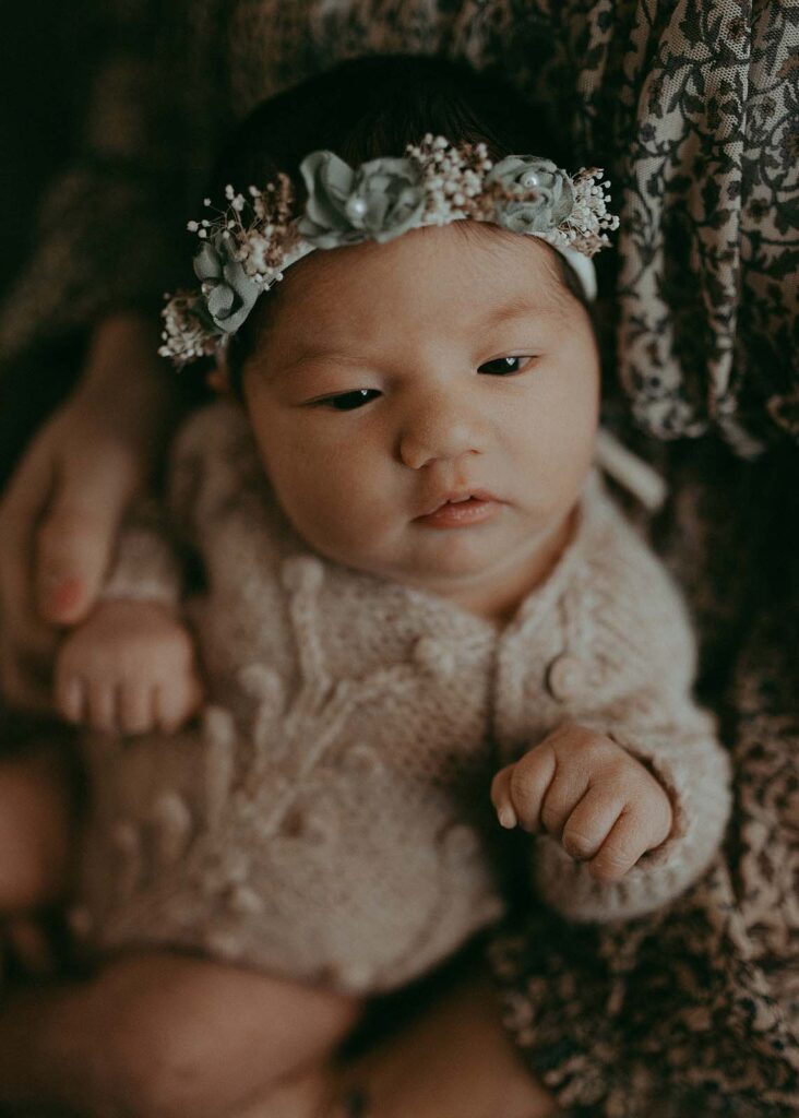 The portrait of newborn girl in a knitted tan romper and flower halo in green tones in mom's laps. The portrait was taken by Victoria Vasilyeva Photography Duke Perinatal Durham NC