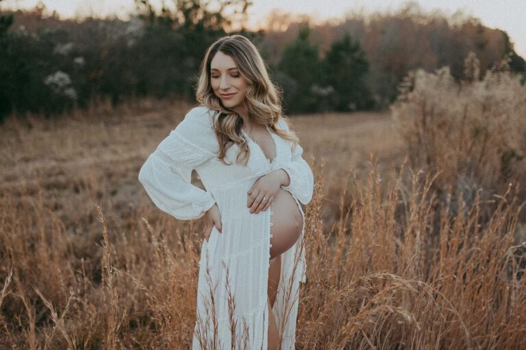 Beautiful mom-to-be surrounded by high grass is stanging in the middle of the field near Oh Baby 4D Ultrasounds in Clayton, NC. The portrait was taken by -Victoria Vasilyeva Photography - Clayton newborn photographer.
