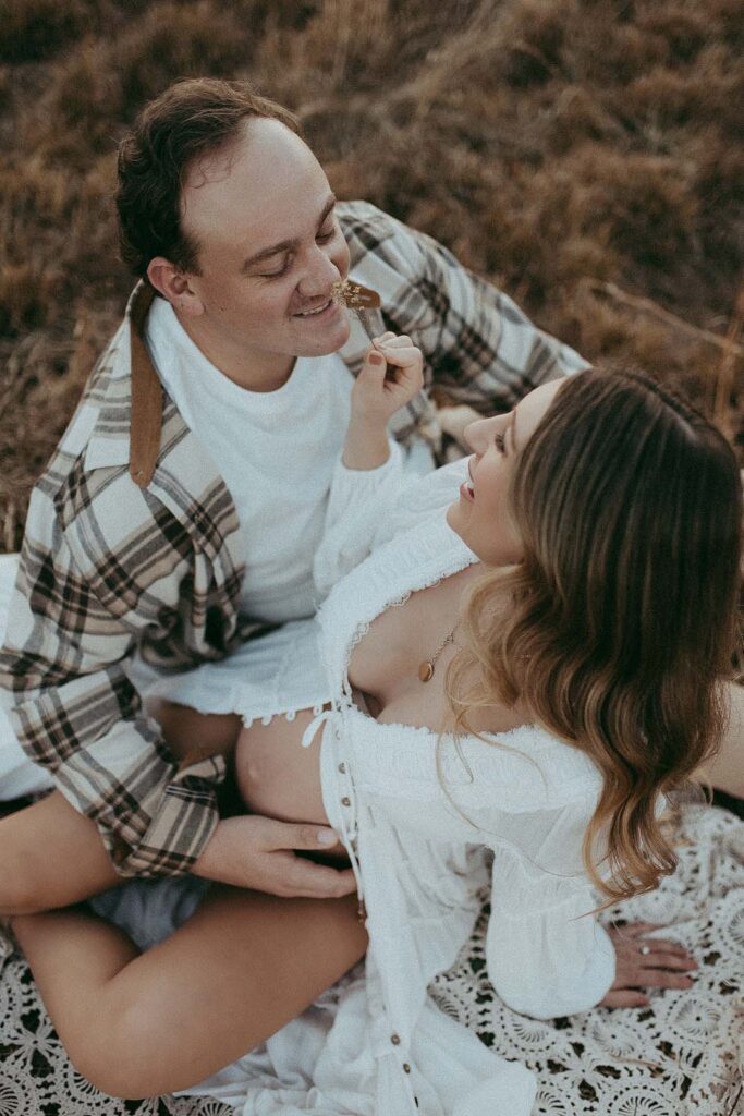 Mom in boho white robe from free people brand and dad in plaid shirt are sitting on a crochet blanket. They are smiling and looking at each other and holding the belly oh baby 4D ultrasounds.