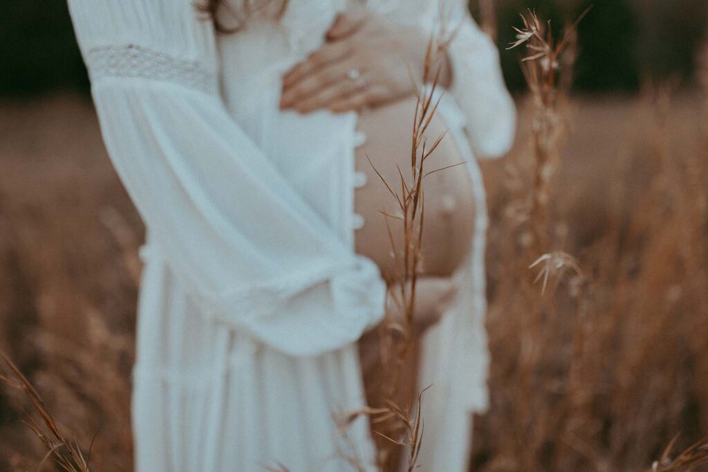 Photo of big belly bump. Mom wears white boho dress and holds her belly tight. The photo was taken among high grass on a field by Victoria Vasilyeva Photography.