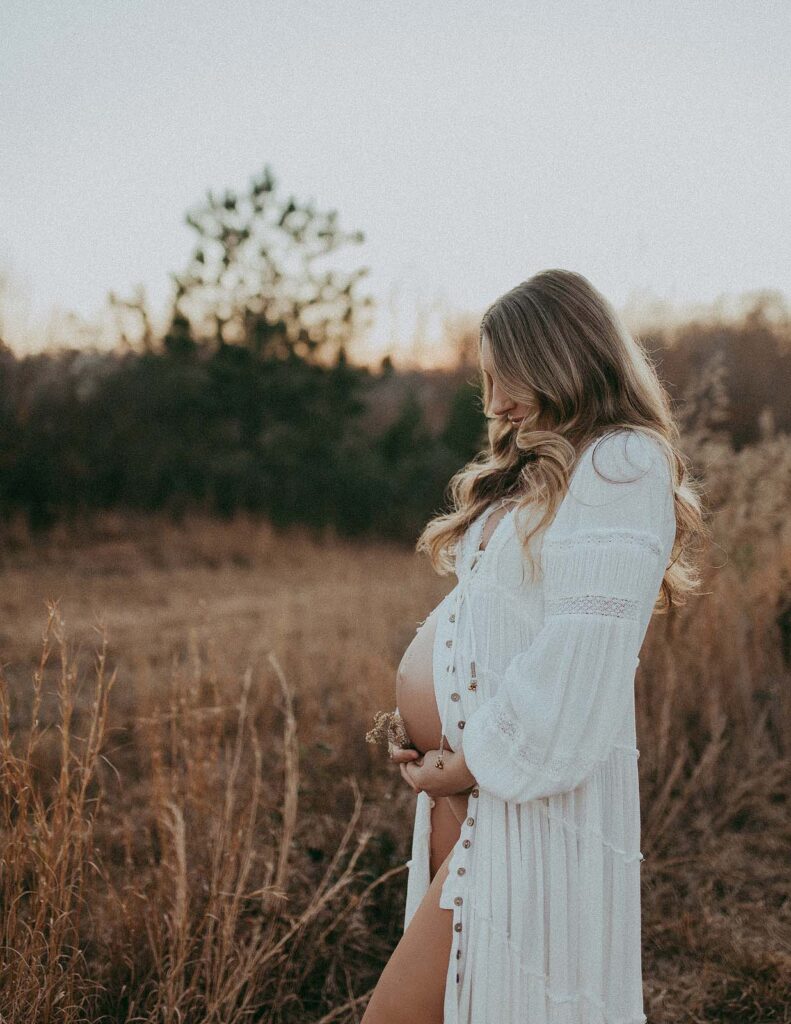 Gorgeous mom-to-be is staying in the middle of the field in white boho dress. She has long wavy hair. Mom is holding the belly and looking down at it. he portrait was taken by maternity photographer - Victoria Vasilyeva Photography near Oh Baby 4D Ultrasounds in Clayton, NC.