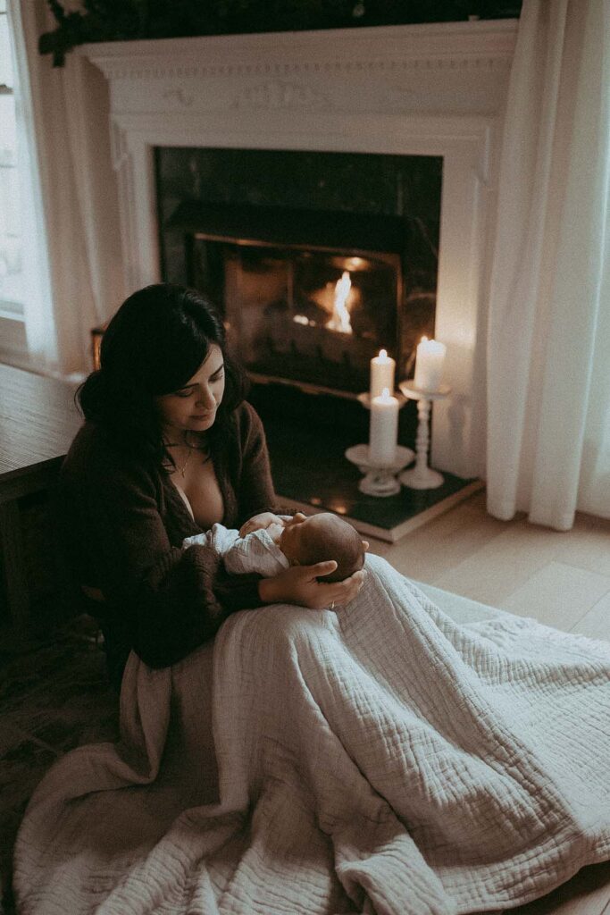 Family portrait of mommy and newborn baby girl. They are sitting on a floor of a living room near the fireplace. Mom's legs covered with a cozy blanket. She is looking down at baby with love and gently hugs her head water birth raleigh nc. Newborn photo session by Victoria Vasilyeva Photography.