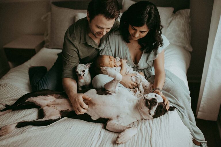 Casual family portra of family with newborn baby girl and 2 puppies. Parents are sitting on a bed, petting the puppy and looking down at her. The family wear cozy clothes in earthy muted tones water birth raleigh nc. Newborn session took place in Raleigh, NC.
