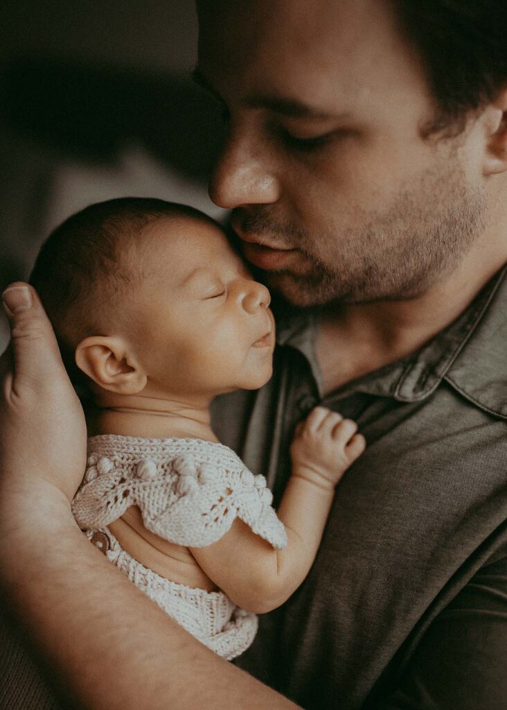 The portrait of Dady and his baby girl. She is sleepy and he kisses her forehead. Baby wears off-white boho romper. The family portrait was taken by Victoria Vasilyeva Photography - Raleigh Newborn Photographer.