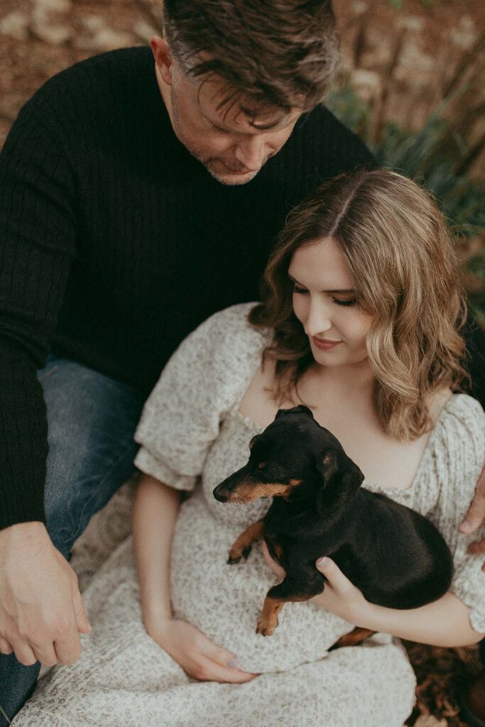 In a tender moment, an anticipating couple shares a seat, the mother-to-be donning an elegant maxi dress while the father-to-be opts for a snug knitted sweater. Their excitement is palpable as they gently hug, a miniature dachshund adding an adorable touch to the scene. The portrait was taken after visiting Barefoot Babies ultrasound studio by Fayetteville maternity photographer - Victoria Vasilyeva Photography.