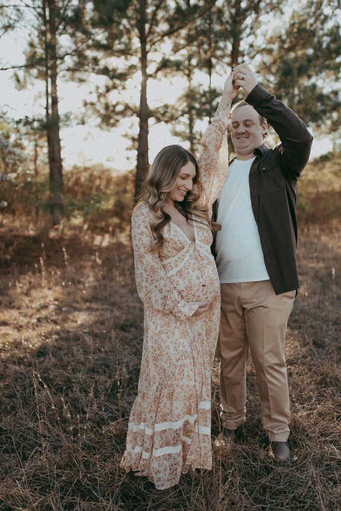 In this heartwarming image, a soon-to-be mom in a blooming floral maxi dress and her partner in a stylish brown shirt and tan pants share a dance, radiating happiness and anticipation for their upcoming journey into parenthood. Maternity portrait was taken by Victoria Vasilyeva Photography prenetal massage cary nc.