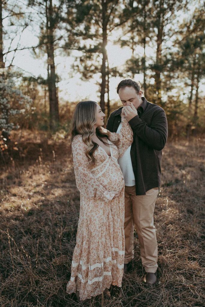 An expecting couple, the mom in a vibrant floral maxi dress, and the dad in a brown shirt and tan pants, share a tender moment as they embrace and dance together. Maternity portrait was taken by Victoria Vasilyeva Photography prenetal massage cary nc.
