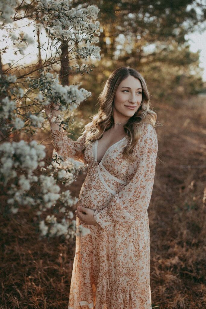 Beautiful mom-to-be in boho maxi dress is staying near blooming bush. She is holding a brunch with her right hand and looking to her left. Maternity photo session took place near Prenatal Massage Cary NC studio by Victoria Vasilyeva Photography.