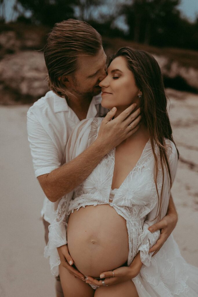 A beautiful expecting couple is sitting on the sand and posing to Victoria Vasilyeva Photography - one of the best maternity and family photographers in Wilmington, NC, and the surrounding area.