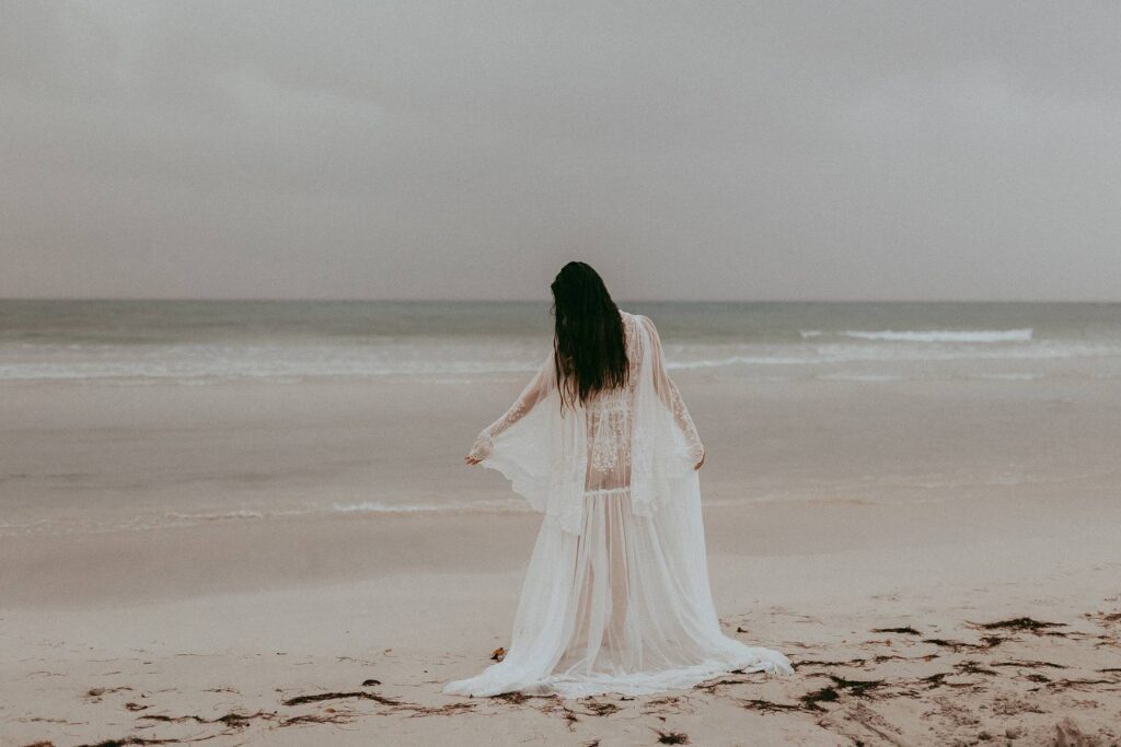 Gorgeous mom-to-be in white lace robe is dancing on a beach during her maternity photo session. Victoria Vasilyeva Photography is one of the best maternity and family photographers in Wilmington, NC