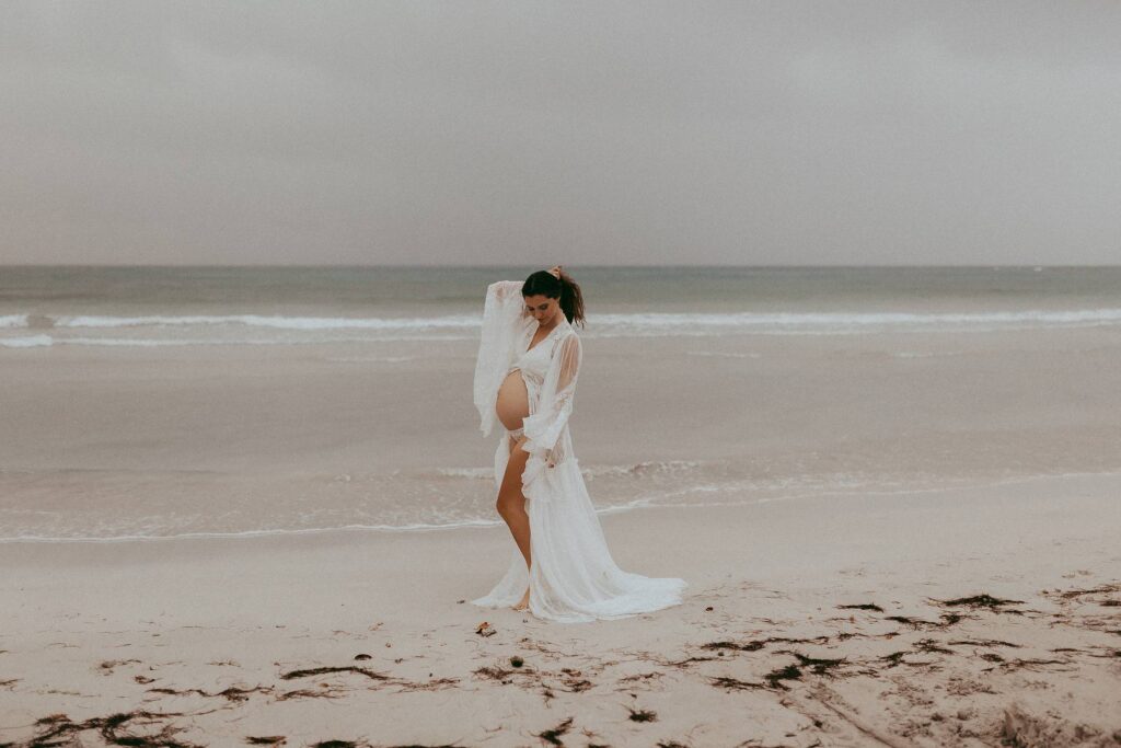 Gorgeous mom-to-be in white lace robe is dancing on a beach during her maternity photo session. Victoria Vasilyeva Photography is one of the best maternity and family photographers in Wilmington, NC