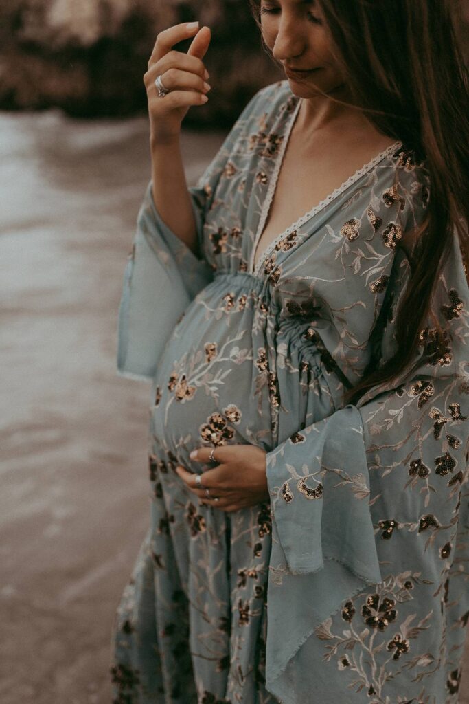 A lovely mom-to-be in a maxi blue dress is standing among rocks and posing to Victoria Vasilyeva Photography - the award-winning Wilmington photographer, specializing in newborn, maternity, and family photographs.