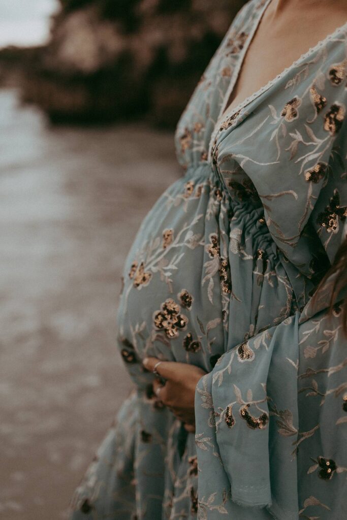 A lovely mom-to-be in a maxi blue dress is standing among rocks and posing to Victoria Vasilyeva Photography - the award-winning Wilmington photographer, specializing in newborn, maternity, and family photographs.