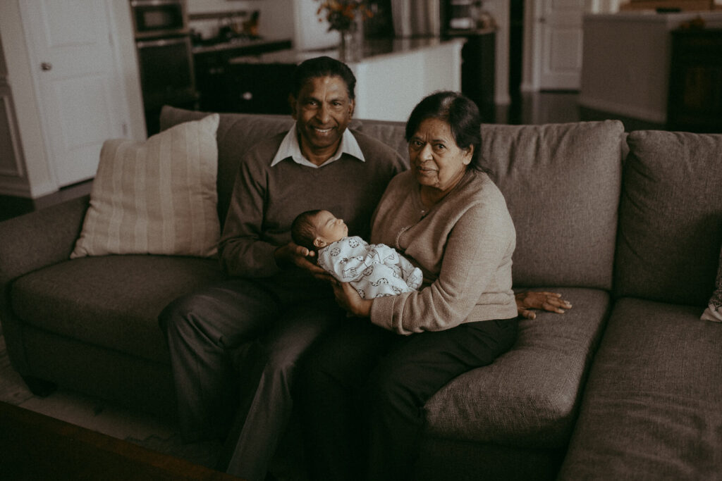 Grandparents with the 4-day-old grandson are sitting on a couch in the living room. Victoria Vasilyeva Photography - Certified, insured, and professional baby photographer in Greensboro. All outfits for baby, mom, and siblings are provided. Newborn Photography Greensboro NC