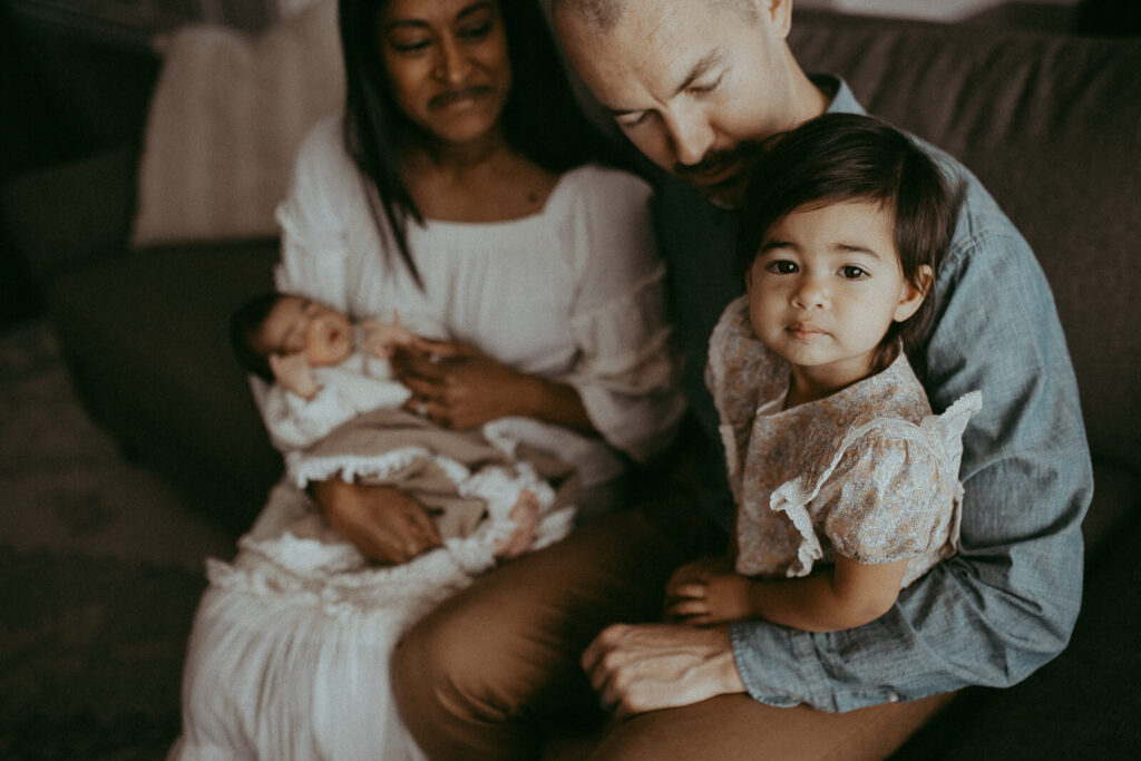 Parents with the 4-day-old son and older daughter are sitting on a couch in the living room. Victoria Vasilyeva Photography - Certified, insured, and professional baby photographer in Greensboro. All outfits for baby, mom, and siblings are provided. Newborn Photography Greensboro NC