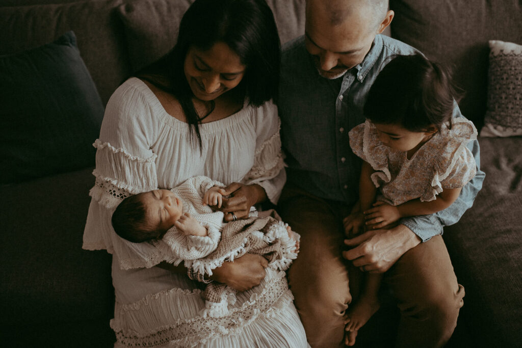Parents with the 4-day-old son and older daughter are sitting on a couch in the living room. Victoria Vasilyeva Photography - Certified, insured, and professional baby photographer in Greensboro. All outfits for baby, mom, and siblings are provided.