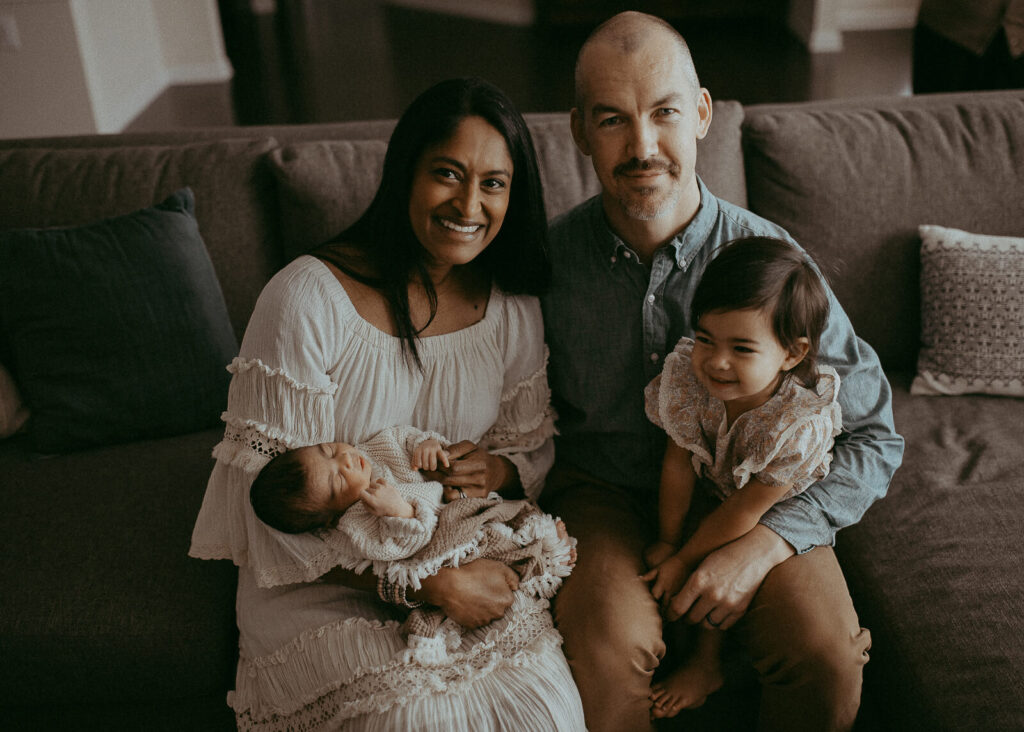 Happy parents with the 4-day-old son and older daughter are sitting on a couch in the living room. Victoria Vasilyeva Photography - Certified, insured, and professional baby photographer in Greensboro. All outfits for baby, mom, and siblings are provided. Newborn Photography Greensboro NC