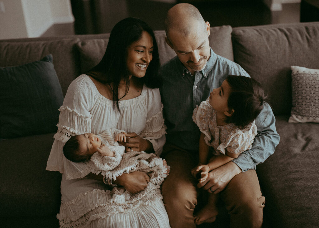 Beautiful family is sitting on a couch in the living room. Victoria Vasilyeva Photography - Certified, insured, and professional baby photographer in Greensboro. All outfits for baby, mom, and siblings are provided. Newborn Photography Greensboro NC