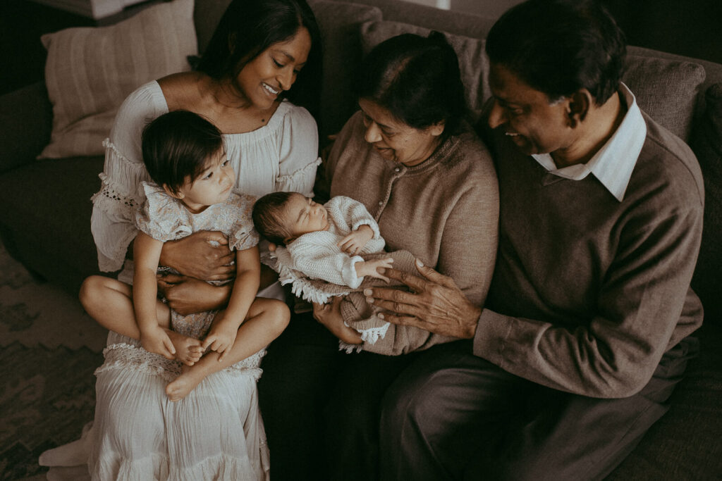 Mom, grandparents with the 4-day-old baby boy and older sibling are sitting on a couch in the living room. Victoria Vasilyeva Photography - Certified, insured, and professional newborn and family photographer in Greensboro. All outfits for baby, mom, and siblings are provided.