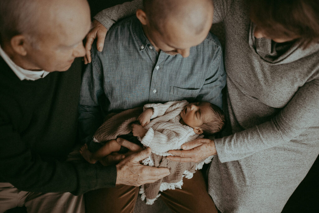 Dad, grandparents with the 4-day-old baby boy are sitting on a couch in the living room. Victoria Vasilyeva Photography - Certified, insured, and professional baby photographer in Greensboro. All outfits for baby, mom, and siblings are provided. Newborn Photography Greensboro NC