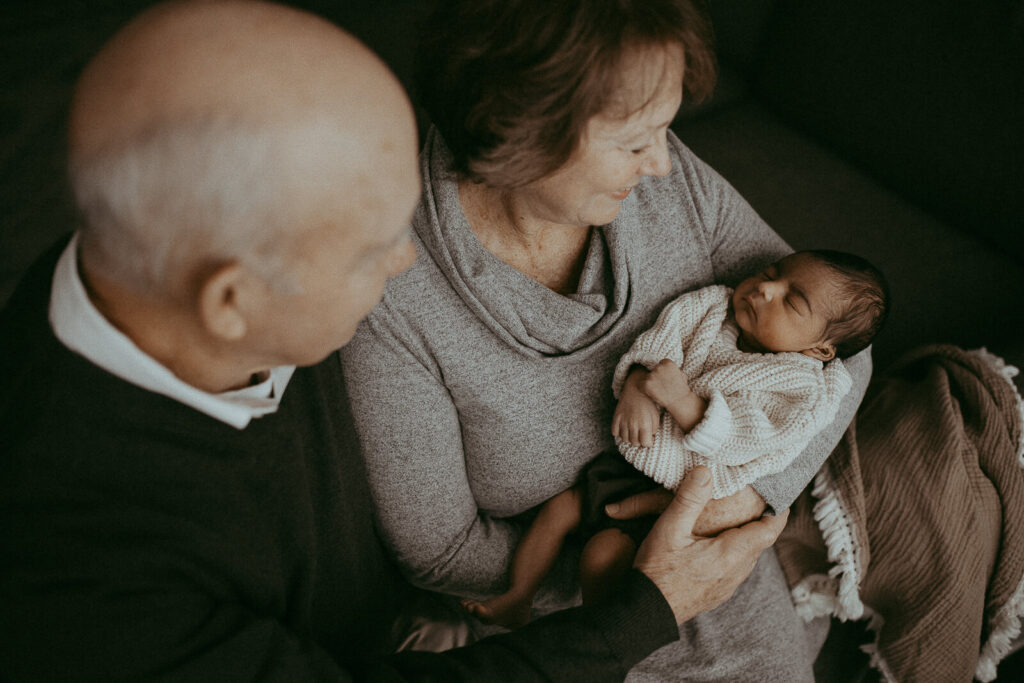 Grandparents with the 4-day-old grandson are sitting on a couch in the living room. Victoria Vasilyeva Photography - Certified, insured, and professional baby photographer in Greensboro. All outfits for baby, mom, and siblings are provided.