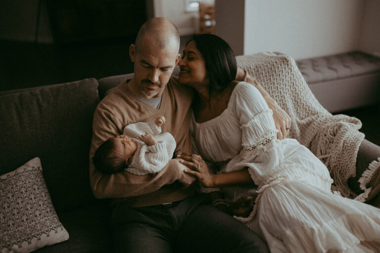 Mom and dad with the 4-day-old son are sitting on a couch in the living room. Mom looks at dad and smiles to him. Victoria Vasilyeva Photography - Certified, insured, and professional baby photographer in Greensboro. All outfits for baby, mom, and siblings are provided.