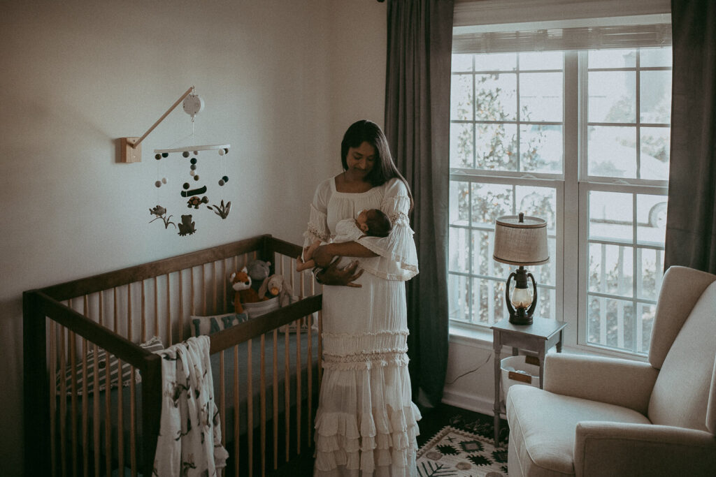 4-days-old baby boy and his mom in white boho dress are in nursery. The portrait was taken by Victoria Vasilyeva Photography - Certified, insured, and professional newborn photographer in Greensboro. All outfits for baby, mom, and siblings are provided.