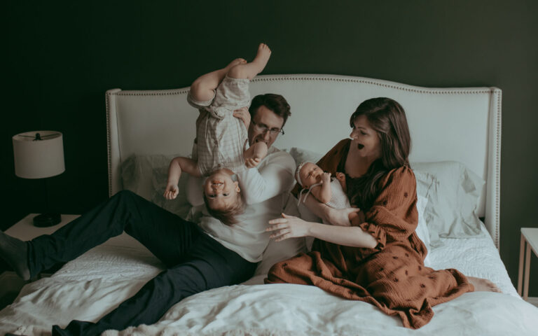 Mom, dad, and 2 kids are in the master bedroom. Mom wears boho brown dress. Dad is playng with older child are looking at him. They are laughing and posing to a family photographer - Victoria Vasilyeva Photography. Photo session took place near Wake Forest Montessori.
