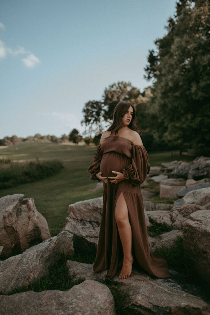 The expecting mother poses in front of huge rocks at NCMA wearing a maxi dress, looking down. Raleigh maternity photographer highlights the natural beauty.