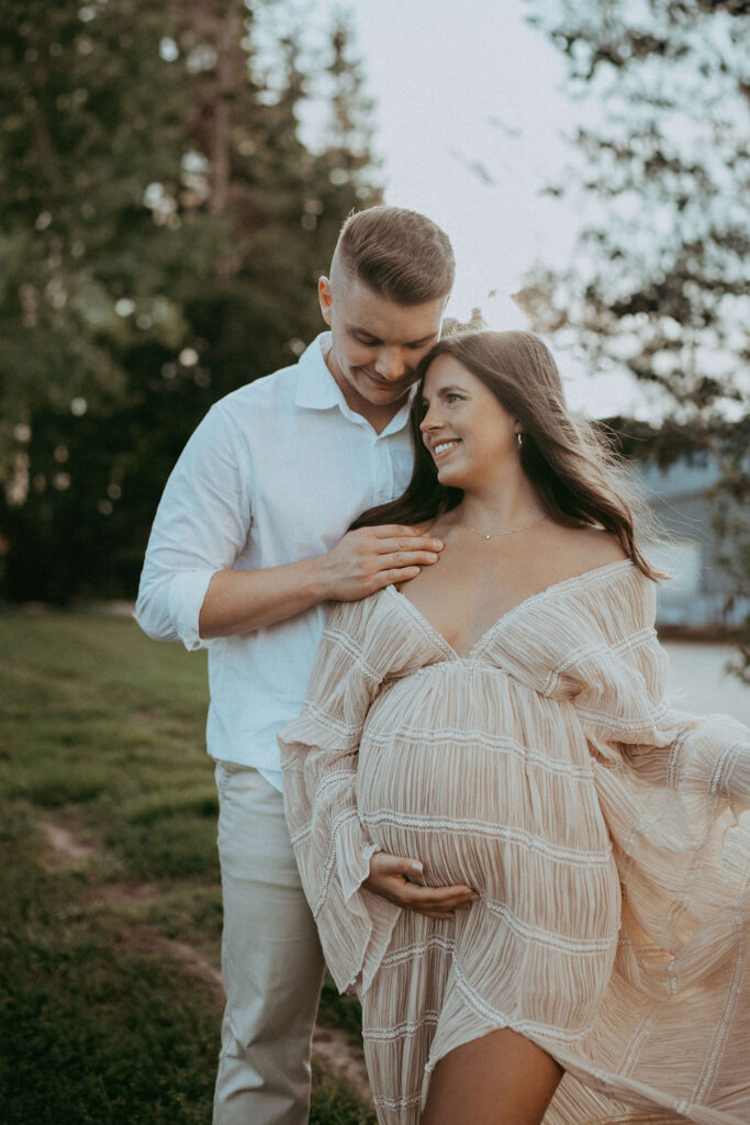 The expecting couple stands together among rocks at NCMA, laughing together. The mom-to-be wears a nude maxi dress, and they share a tender moment. Maternity photo session at NCMA in Raleigh, NC by Victoria Vasilyeva Photography.