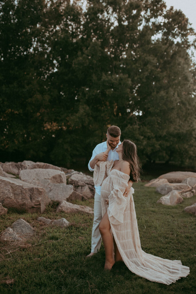 The expecting couple stands together among rocks at NCMA, laughing together. The mom-to-be wears a nude maxi dress, and they share a tender moment. Maternity photoshoot in Raleigh, NC